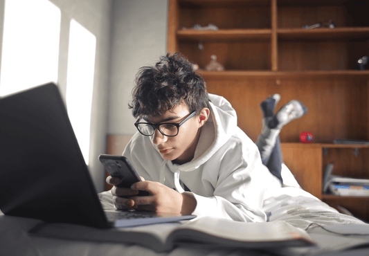 Male teenager with glasses studying and using his phone on the bed